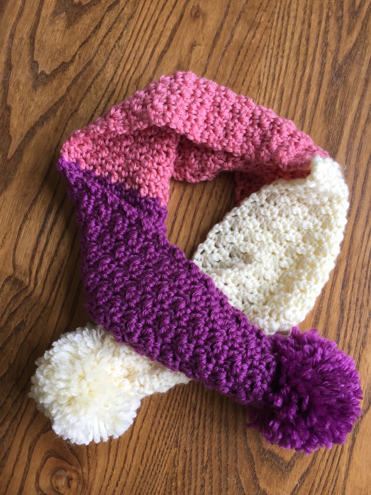 a kids' scarf in purple. pink and white with matching pom poms on each end