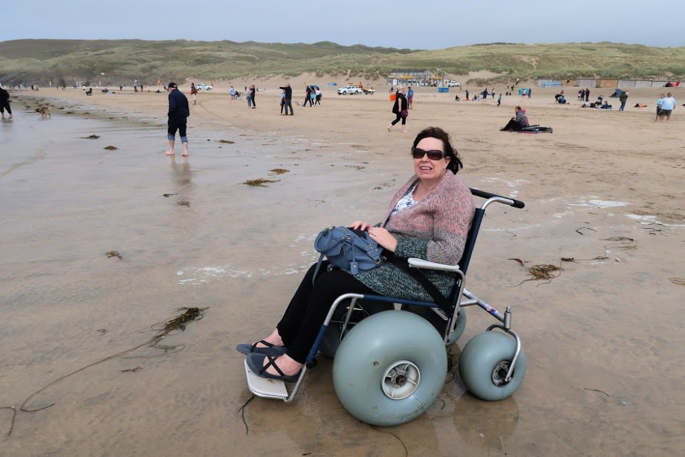 Me in a wheelchair on the beach at Perranporth, Cornwall. The beach has people in the background. The beach wheelchair has big bouncy wheels ,