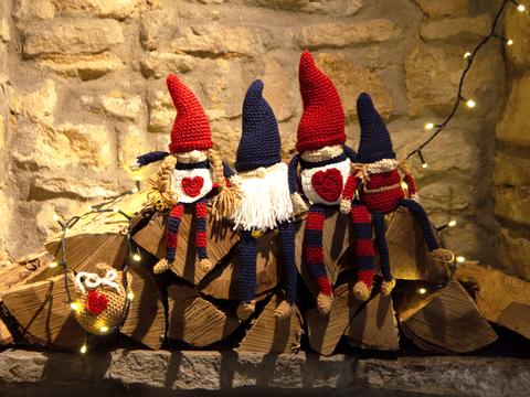 Four Christmas crochet gnomes in blue and red sitting on logs against a brick wall