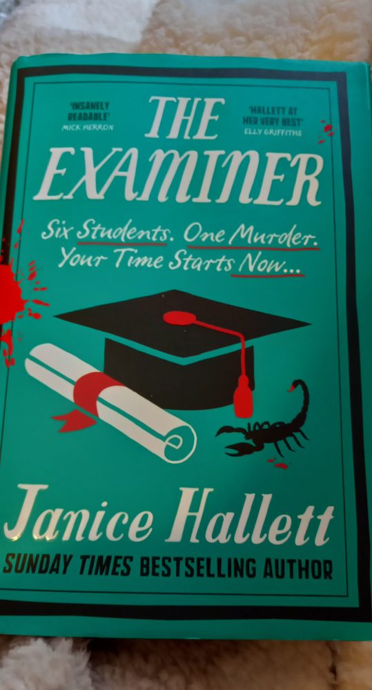 The examiner book cover, image shows a graduate hat, a scroll, a scorpion and it's splattered with blood red blotches. 